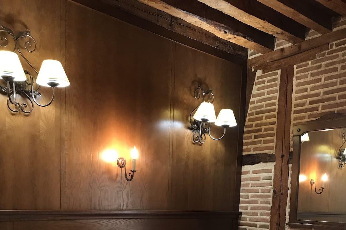 Quarto de hotel com cabeceira de madeira, duas luminárias fixadas na parede, cama com edredom branco, almofada estampada e toalha dobrada com monograma.