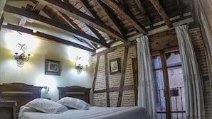 Een kamer met een houten plafond, een bed met een grijze deken, twee wandlampen en een raam met gordijnen.
