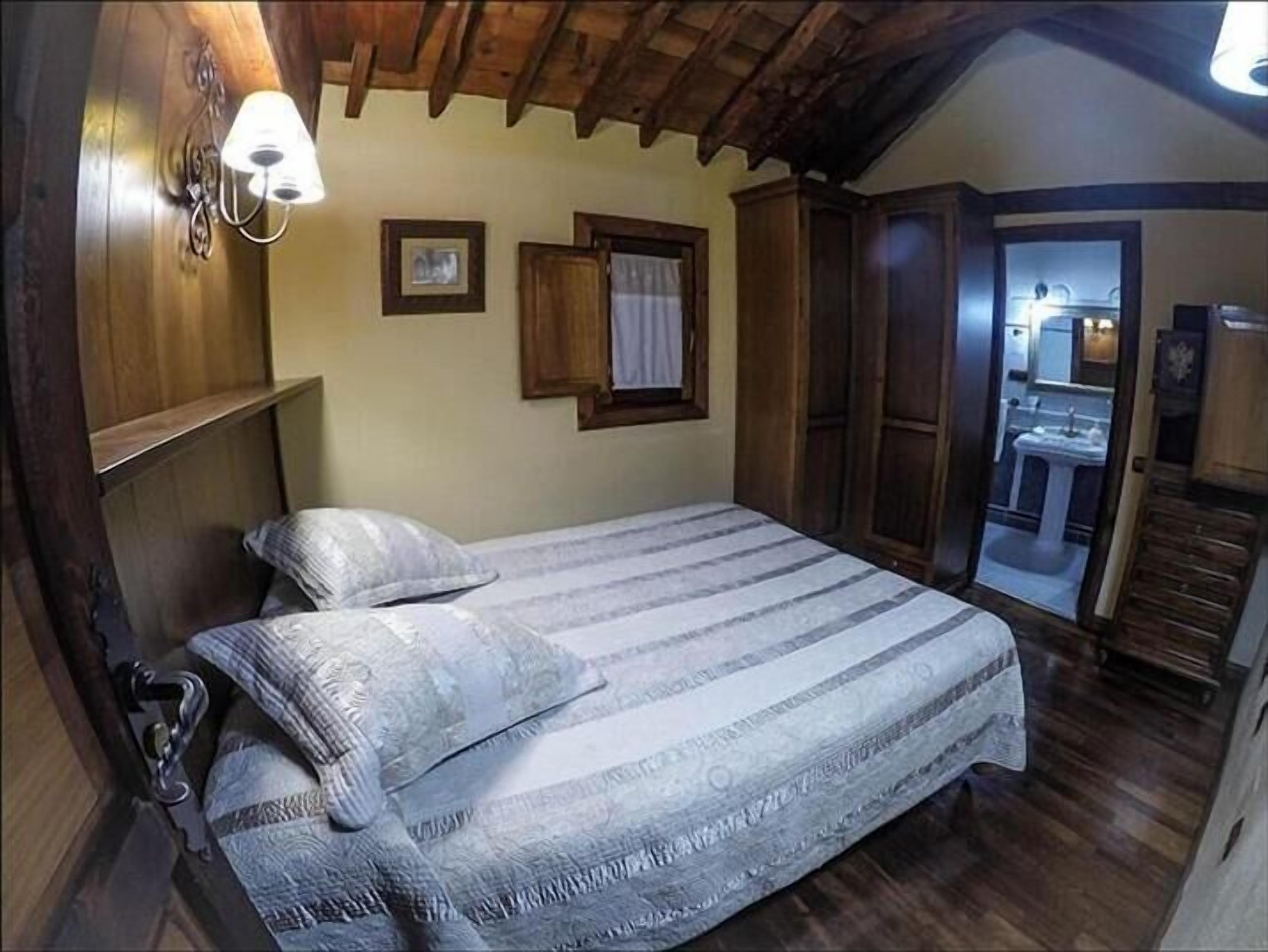 A bedroom with a wooden ceiling, a bed with striped bedding, a nightstand, a wall-mounted lamp, and a door leading to a bathroom.