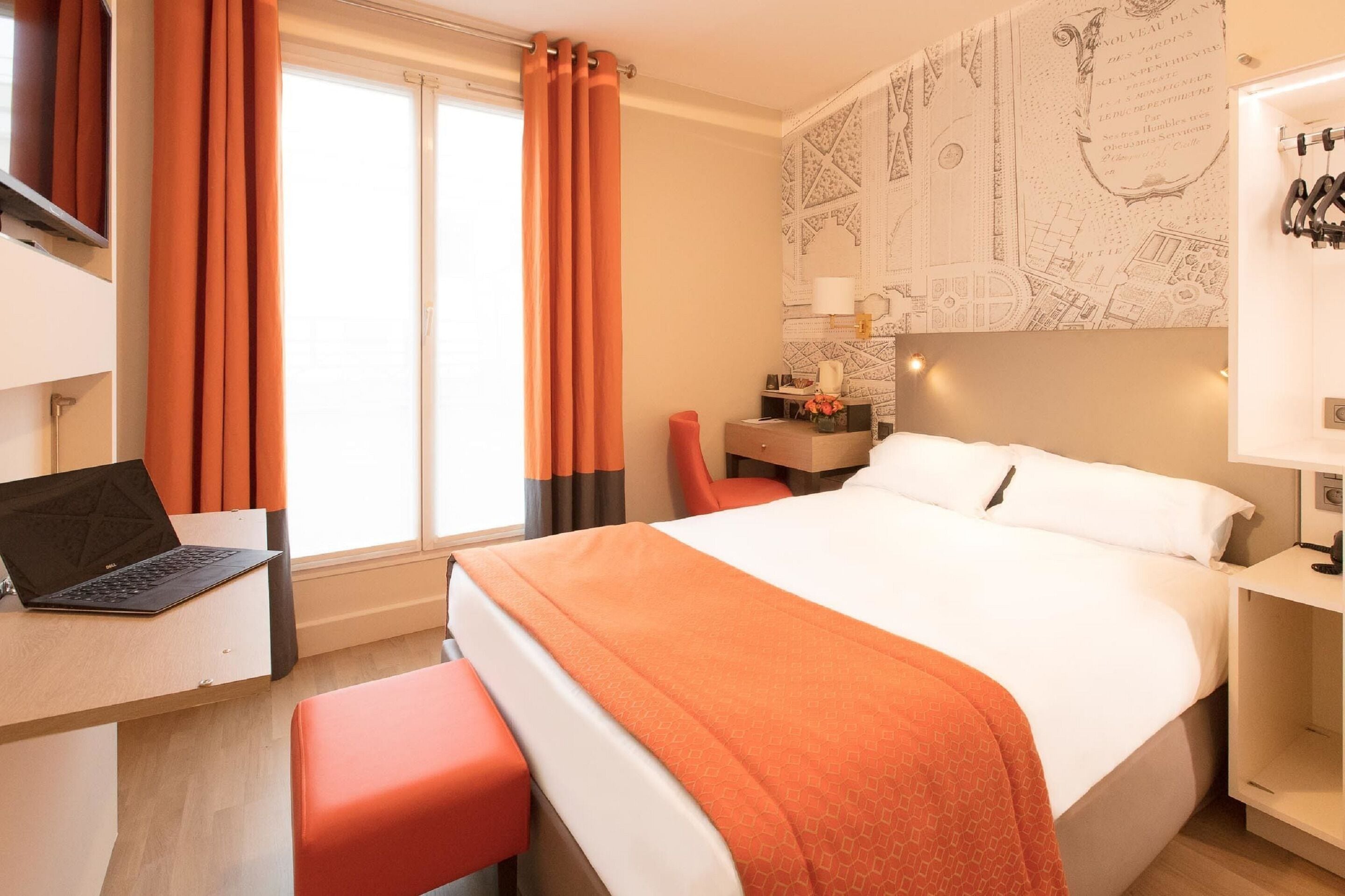 Habitación de hotel con cama, escritorio con computadora portátil, una silla roja y mesita de noche con lámpara.