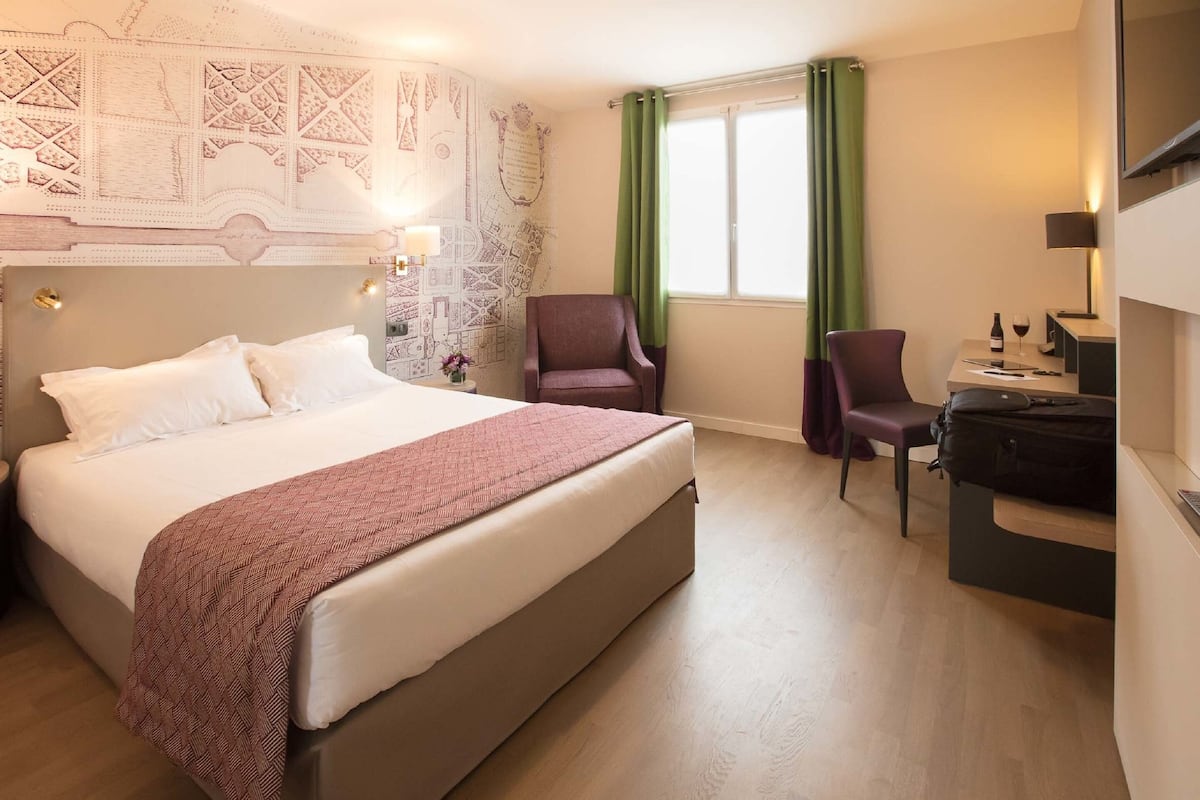 A hotel room with a large bed, two chairs, a desk with a computer, and a window with green curtains.
