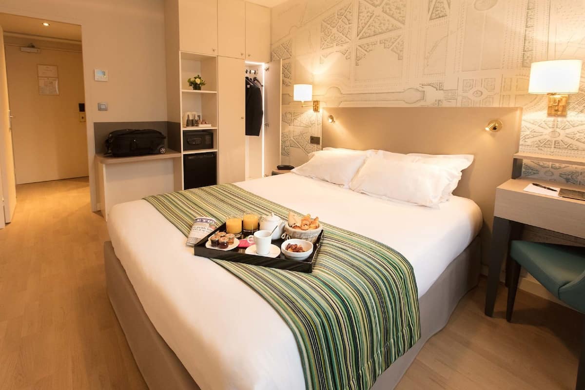 A hotel room with a neatly made bed, a desk, and a tray of breakfast items.