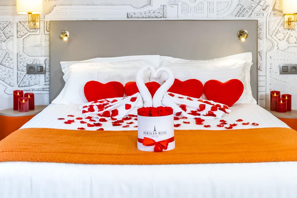 A neatly made bed with white and red heart-shaped pillows, a bouquet of red roses, and a candle on each side.