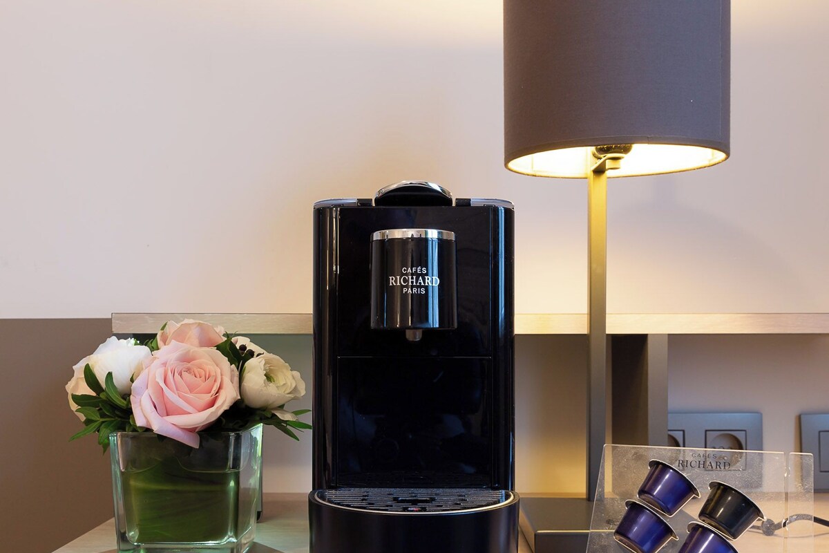 Una cafetera negra con la leyenda 'CAFFES RICHARD PARIS', un florero de vidrio transparente con flores y una lámpara con pantalla oscura sobre una superficie de madera.