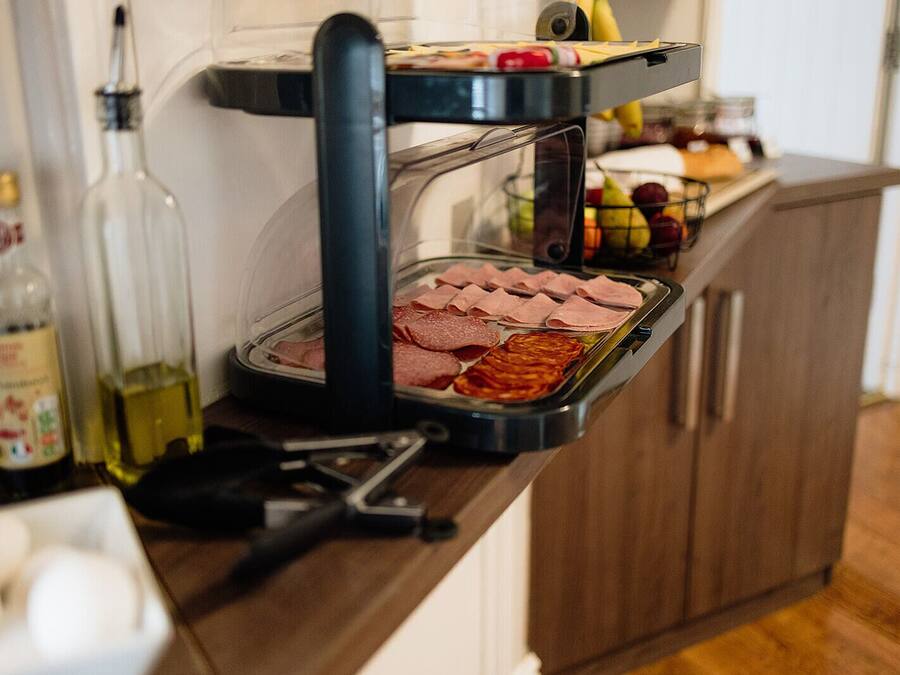 A kitchen counter with a sandwich press containing sliced meats and a fruit basket.