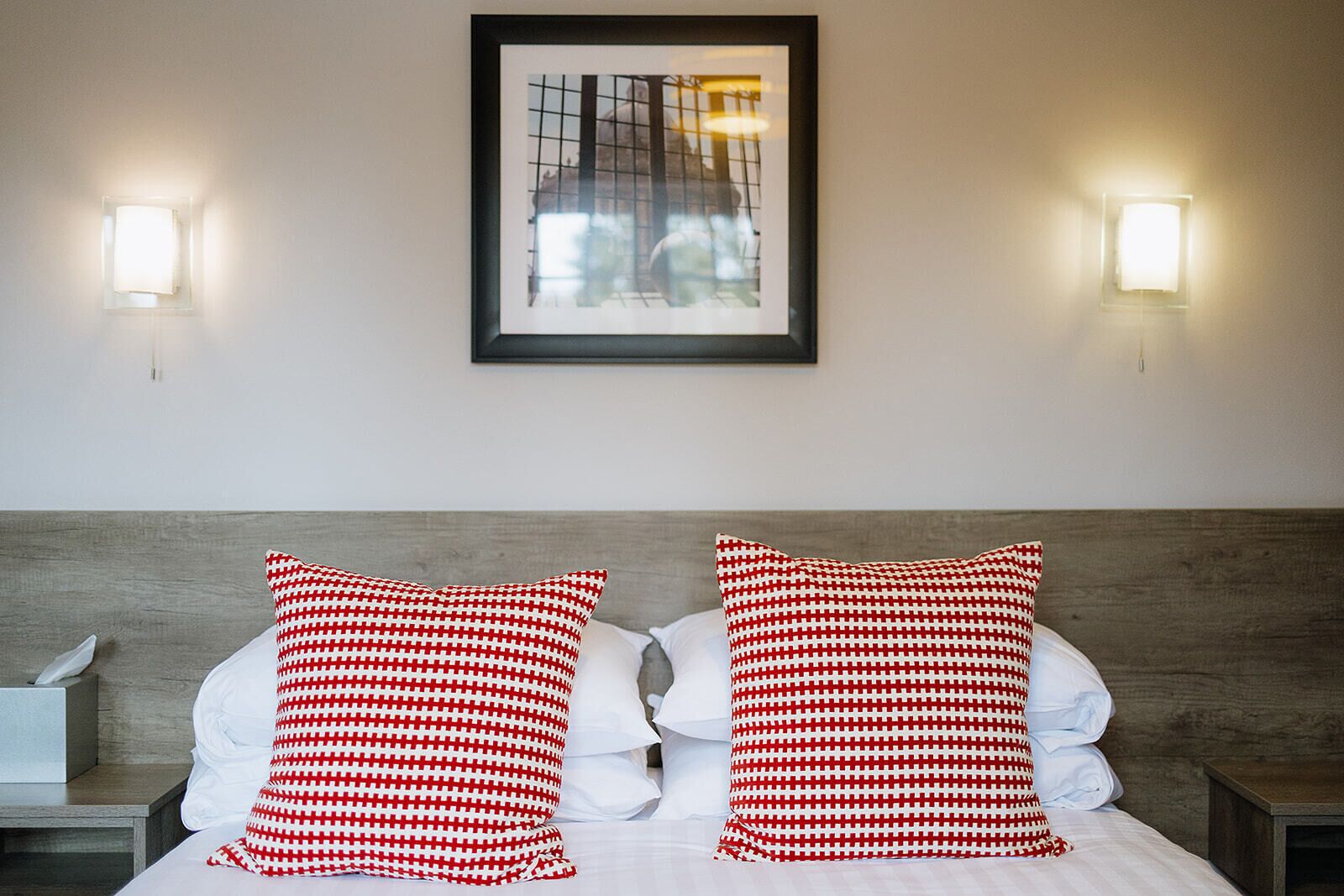 Ein Hotelzimmer mit einem Bett, zwei Kissen mit Karomuster, einem gerahmten Bild an der Wand und zwei Wandleuchten.