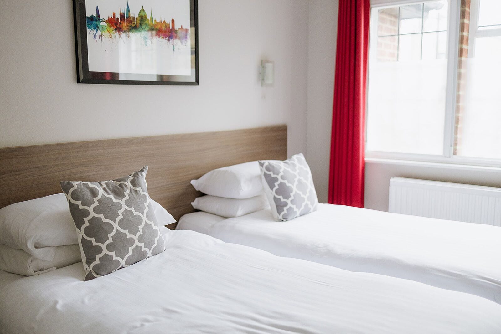 A hotel room with two beds, white bedding, patterned pillows, a framed cityscape artwork, and a window with red curtains.