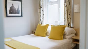 Una cama bien hecha con almohadas amarillas y una colcha blanca, un cuadro en la pared y una ventana con cortinas.