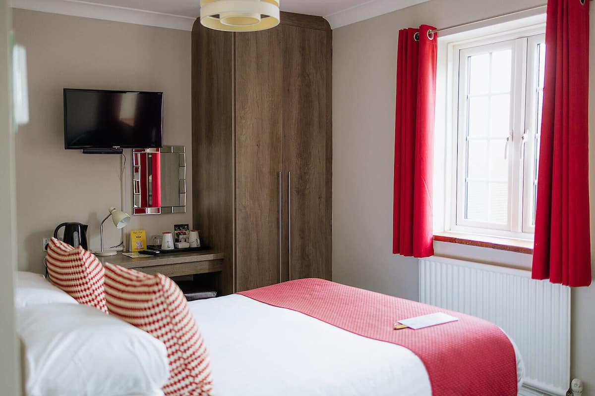 A hotel room with a bed, a desk with a lamp, a television, and a window with red curtains.