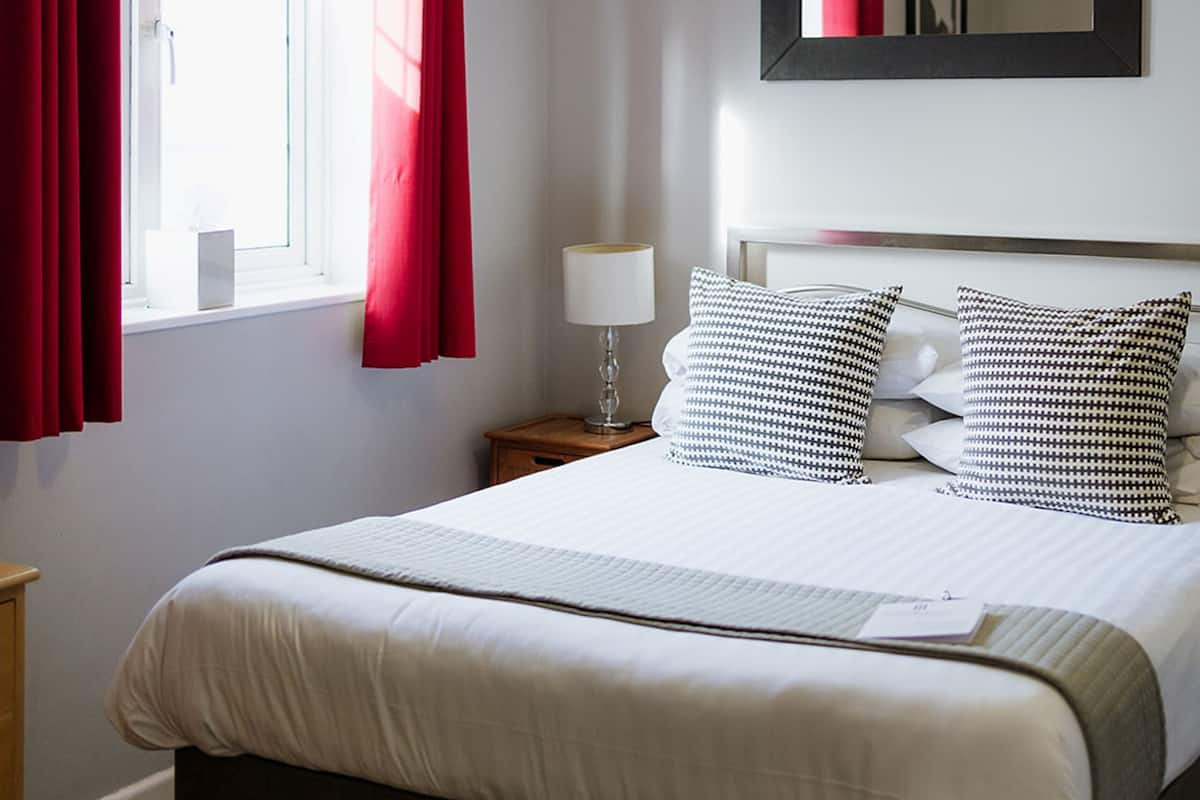 A neatly made bed with white bedding and black and white patterned pillows, a bedside table with a lamp, a window with red curtains, and a hanging light fixture.
