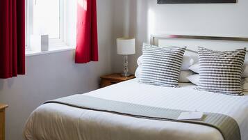 Ein ordentlich bezogenes Bett mit weißer Bettwäsche und Kissen mit schwarz-weißem Muster, ein Nachttisch mit Lampe, ein Fenster mit roten Vorhängen und eine hängende Leuchte.