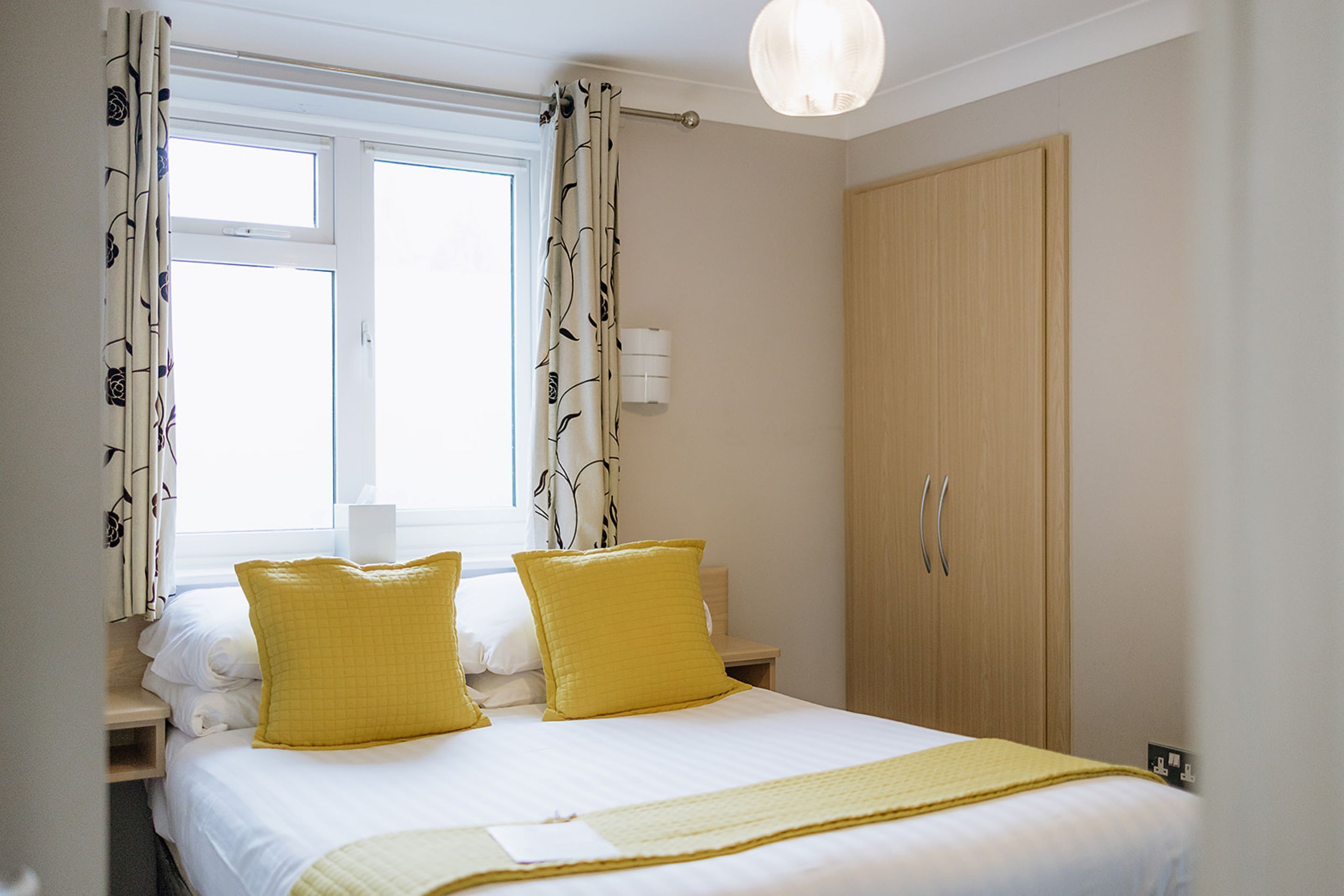 A hotel room with a bed, two yellow pillows, a bedside table with a lamp, a wardrobe, and a window with curtains.