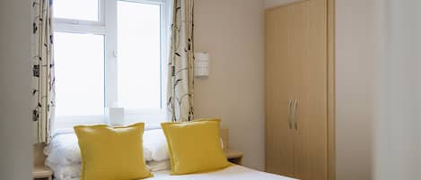 A hotel room with a bed, two yellow pillows, a bedside table with a lamp, a wardrobe, and a window with curtains.