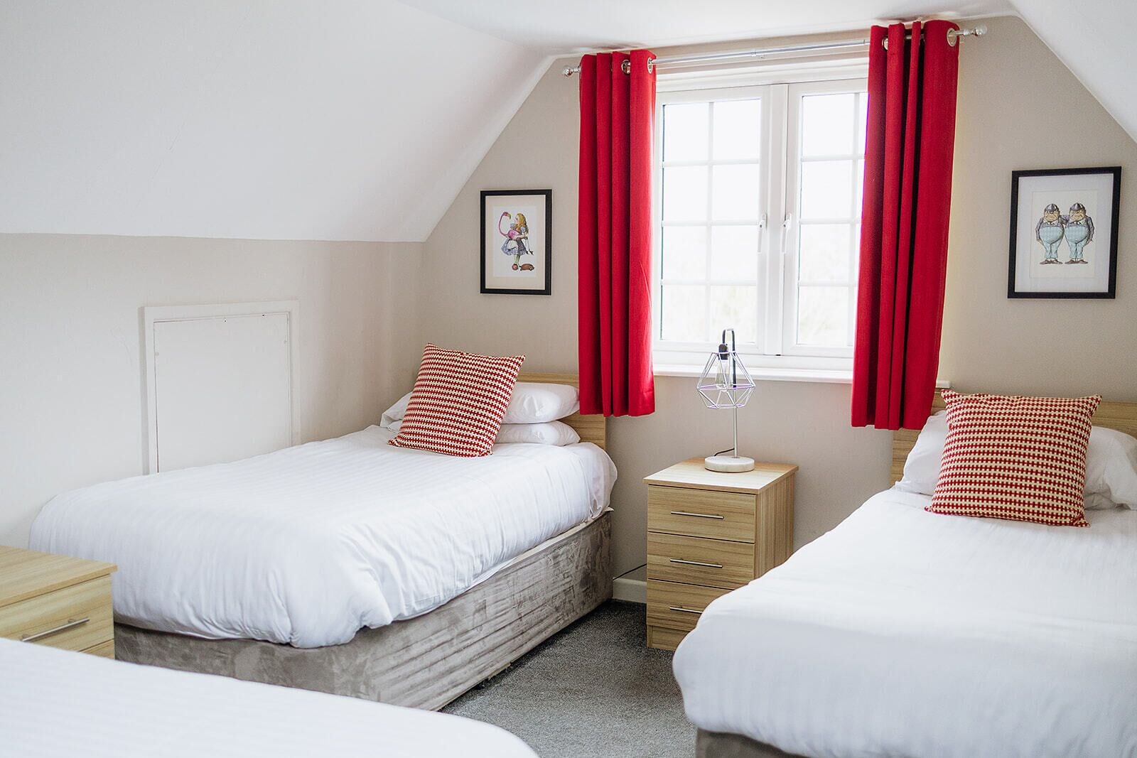 A bedroom with two single beds, a nightstand with a lamp, and a window with red curtains.