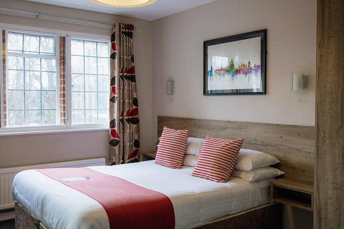 A bedroom with a bed, a window with curtains, a framed cityscape picture, and a bedside table.
