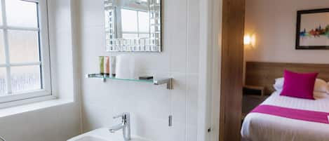 Un baño con un lavabo, repisas de vidrio con artículos de tocador, un espejo y una cama con almohada rosa.