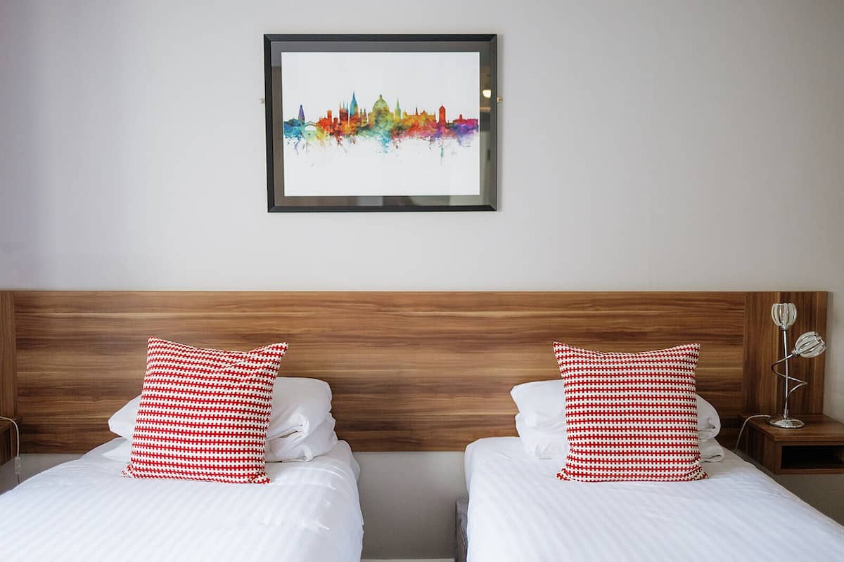 A hotel room with two beds, each with white bedding and red and white checkered pillows. A framed city skyline artwork is on the wall above the beds.