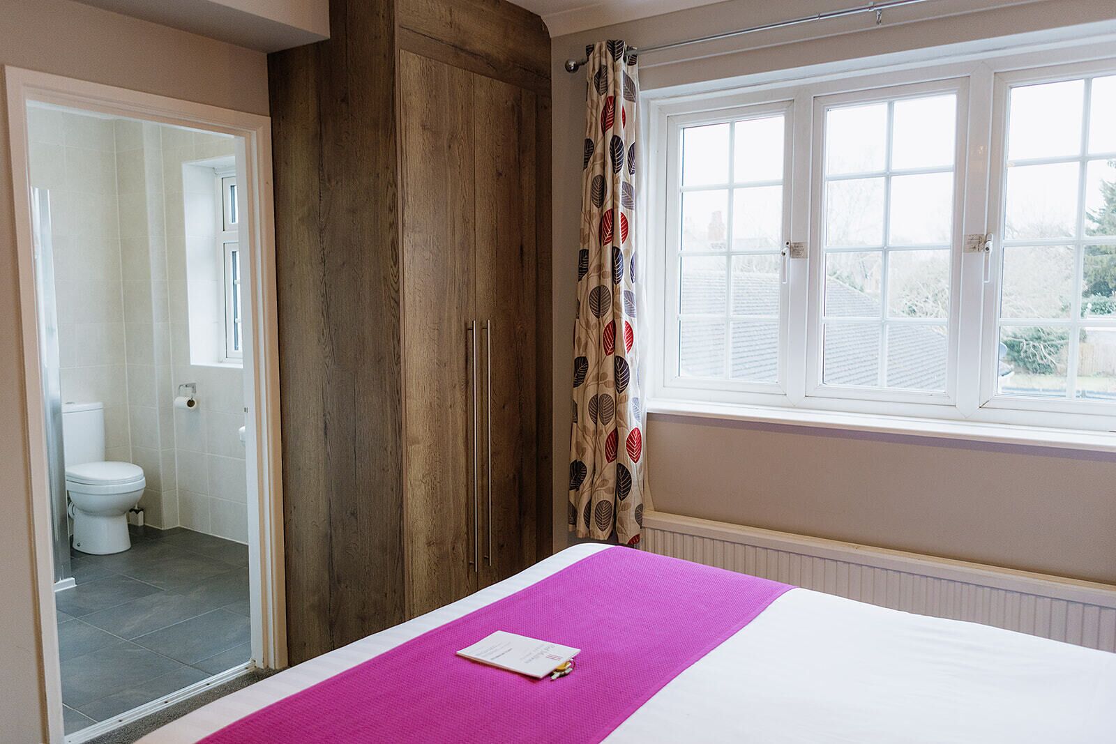 A bedroom with a bed, a window, a wardrobe, and a bathroom visible through an open door.