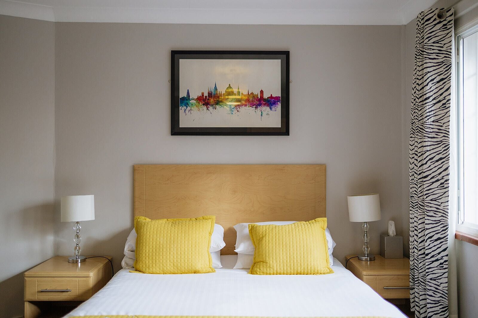A hotel room with a bed, two bedside lamps, a framed cityscape artwork, and zebra-patterned curtains.