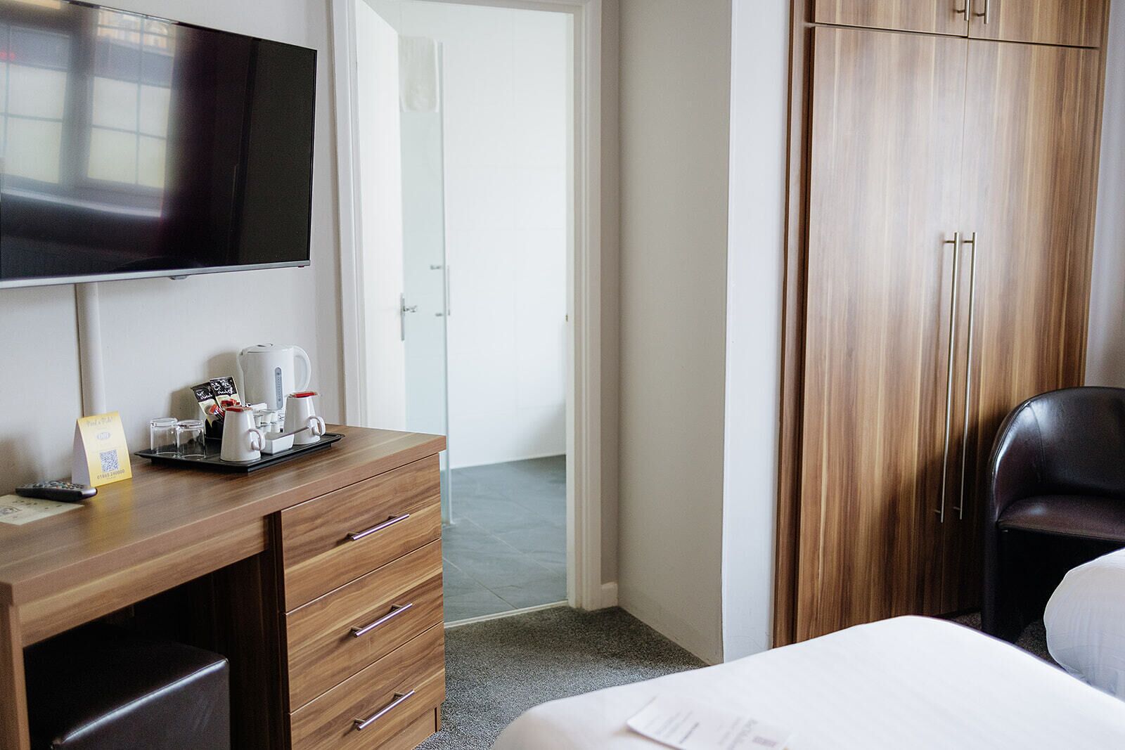 A hotel room with a wooden desk, a flat-screen TV, a bed, and a wardrobe.