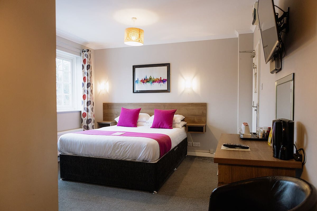 A hotel room with a bed, bedside table, desk, chair, and a large window with curtains.
