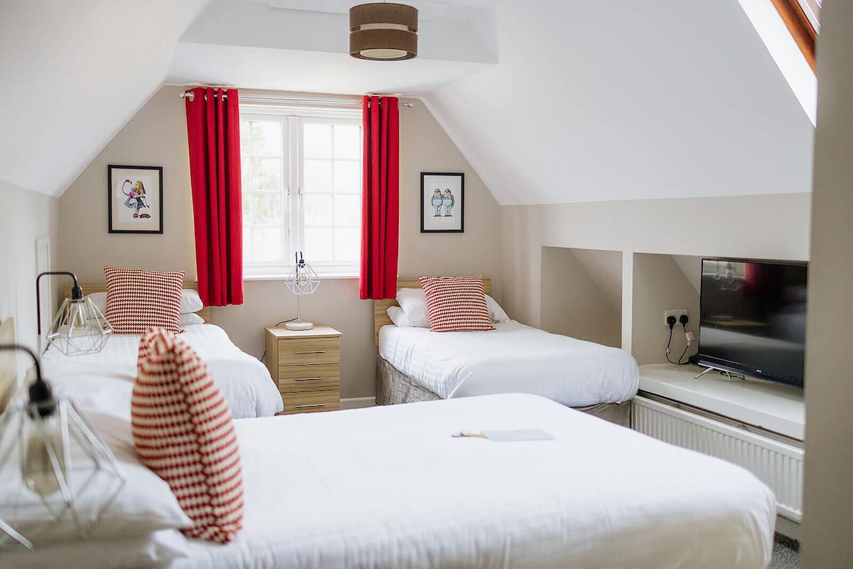 A hotel room with two beds, a bedside table, a lamp, a TV, and a window with red curtains.