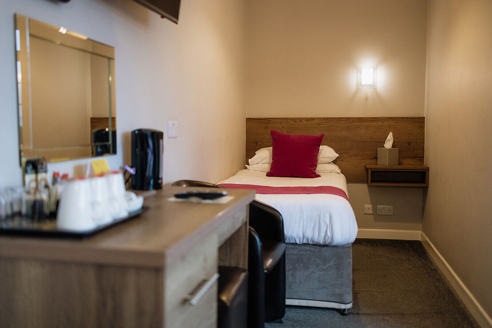A hotel room with a bed, a desk with a coffee maker, a mirror, and a nightstand with a lamp.