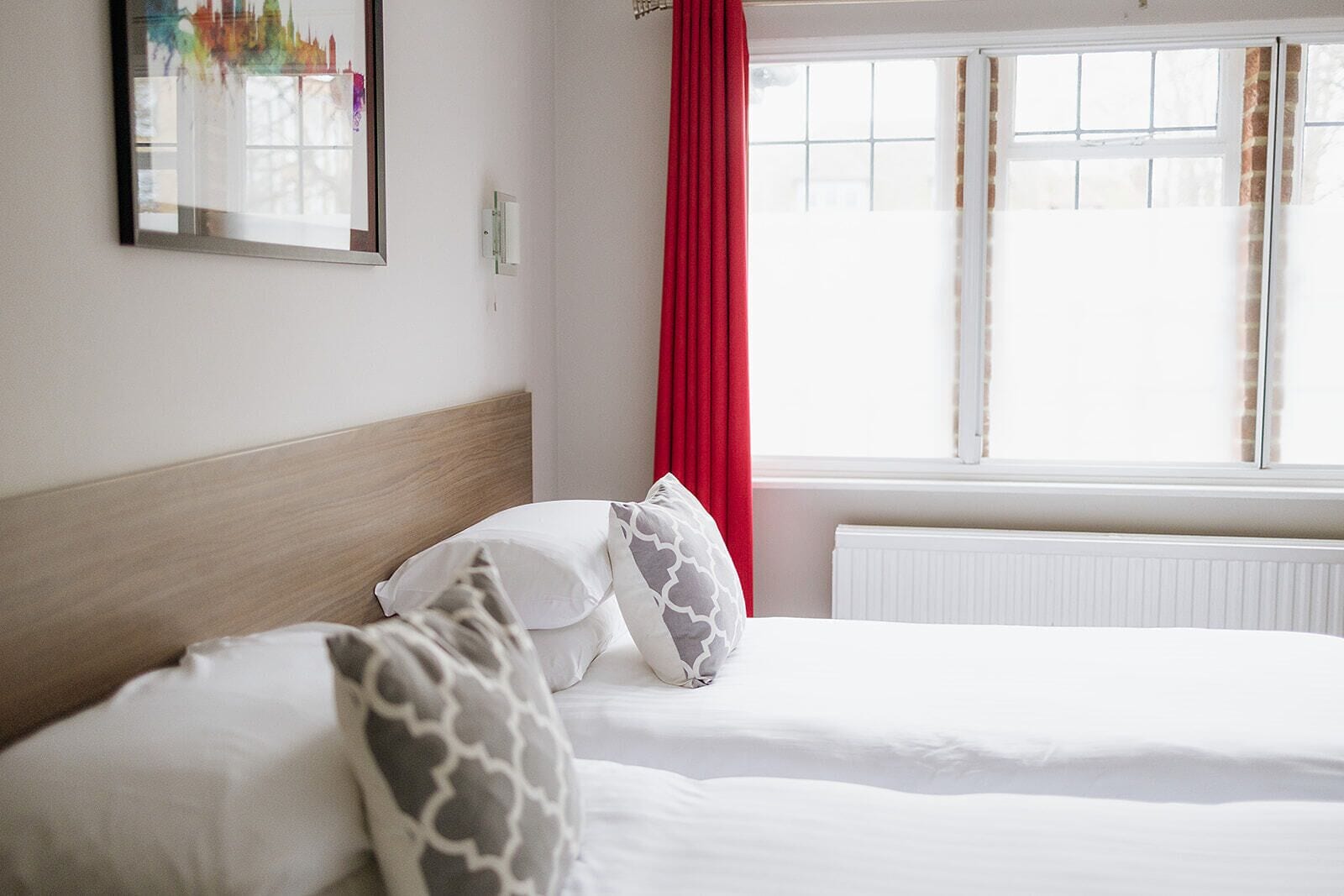 Habitación de hotel con dos camas, cabecero de madera, ventana con cortinas rojas y un cuadro enmarcado en la pared.