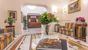 A lobby with a marble floor, a sofa, a coffee table with a book, a vase with flowers, and framed pictures on the walls. - Raeli Hotel Lux (Rome)