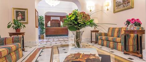 A lobby with a marble floor, a sofa, a coffee table with a book, a vase with flowers, and framed pictures on the walls.