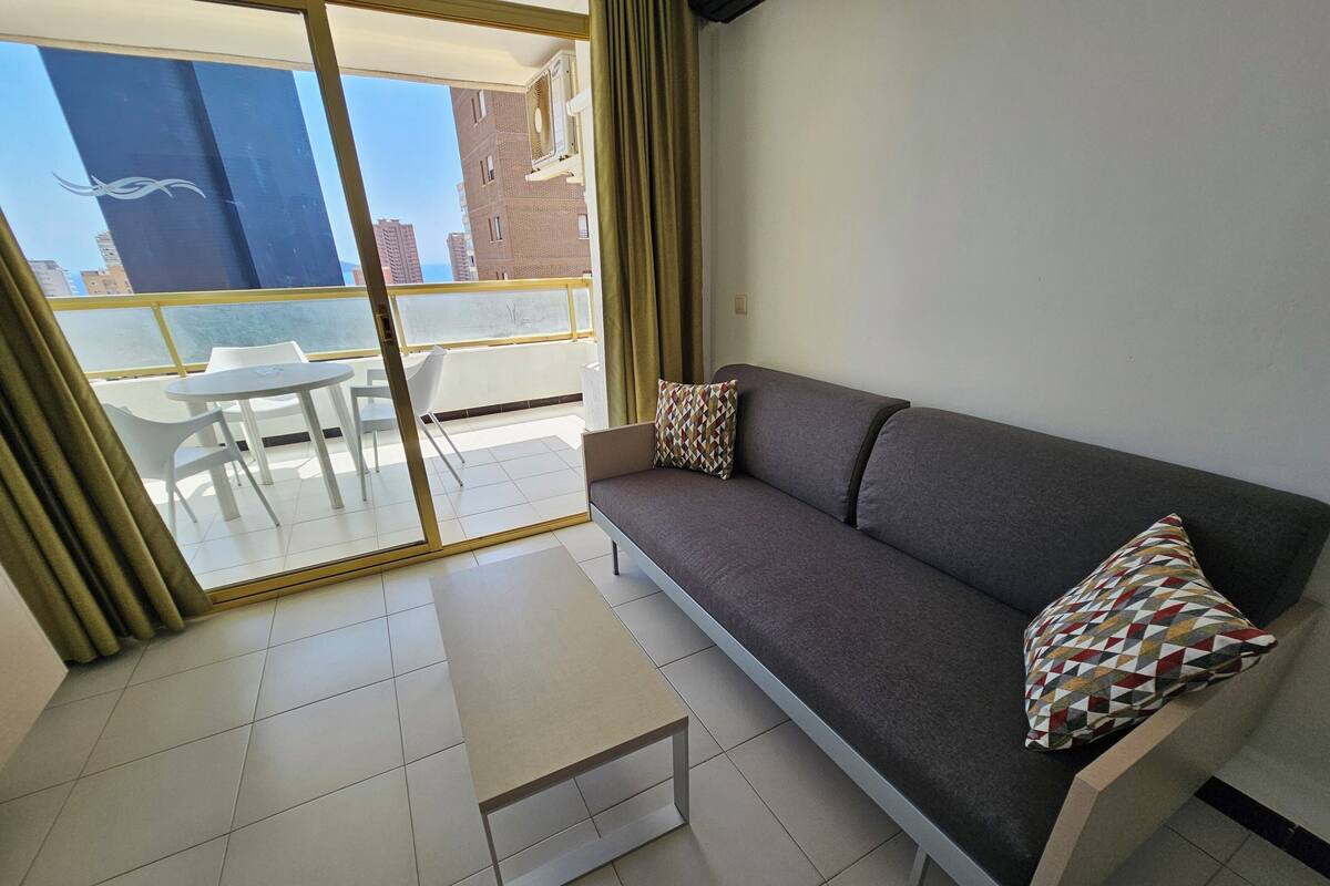 A modern living room with a grey sofa, patterned cushions, a small white table, and a view of a balcony with a glass door.