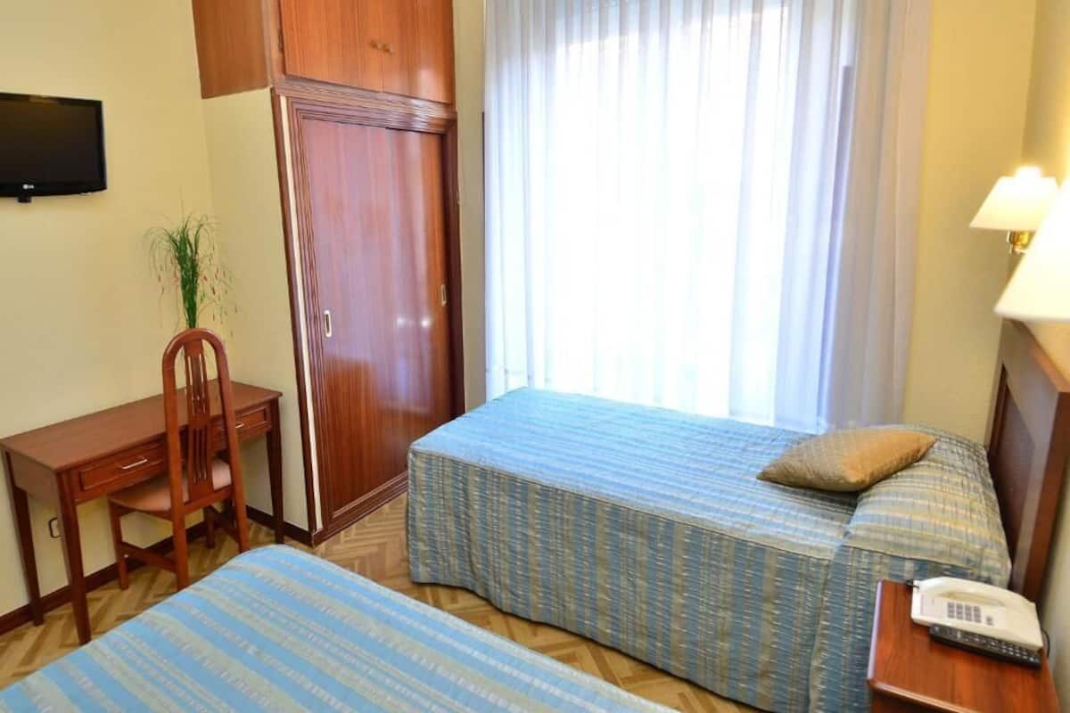 A hotel room with two beds, a desk, a chair, a TV, and a lamp.