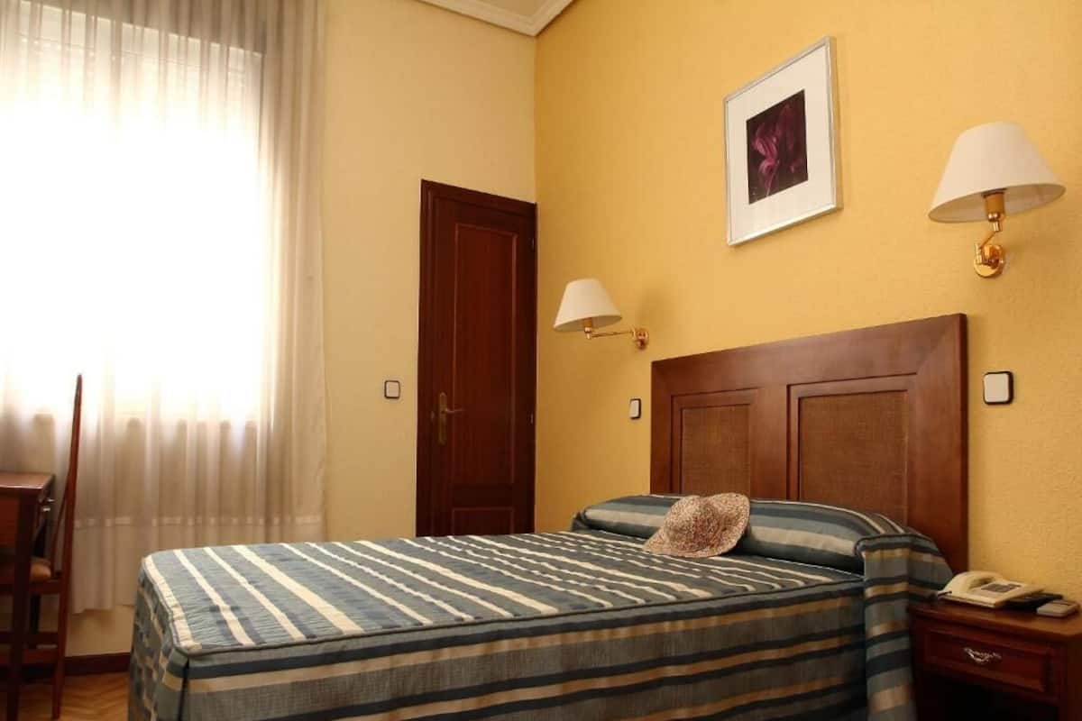 A hotel room with a bed, a nightstand, a chair, a desk, and a window with curtains.
