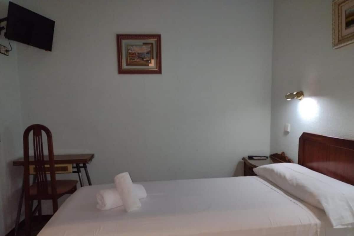 A neatly made bed with white linens, a wooden chair, a small table, a framed picture on the wall, and a bedside lamp.
