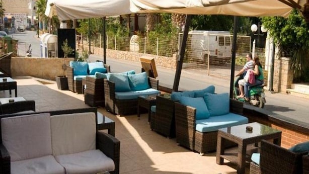 An outdoor seating area with wicker furniture and blue cushions, a covered section with a white canopy, and a view of the street with a parked vehicle and a person on a scooter. - Anemi Apartments (Hersonissos)