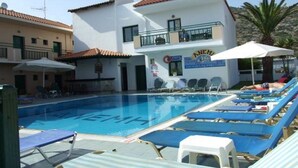 A swimming pool area with lounge chairs and umbrellas, a building with a sign reading "ANEMH", and a clear sky. - Anemi Apartments (Hersonissos)