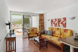 Een woonkamer met een bank, fauteuils, een salontafel en een televisie. Er is een schuifdeur die leidt naar een buitenruimte met een tafel en stoelen.