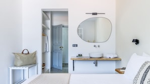 A bedroom with a bed, a bedside table, a mirror, a wooden shelf with toiletries, and a door leading to a bathroom.