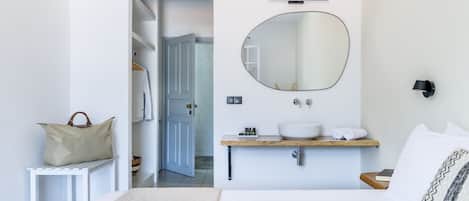 A bedroom with a bed, a bedside table, a mirror, a wooden shelf with toiletries, and a door leading to a bathroom.
