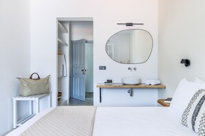 A bedroom with a bed, a bedside table, a mirror, a bedside lamp, a wooden shelf with toiletries, and a door leading to a bathroom. - Aperitton (Skopelos)