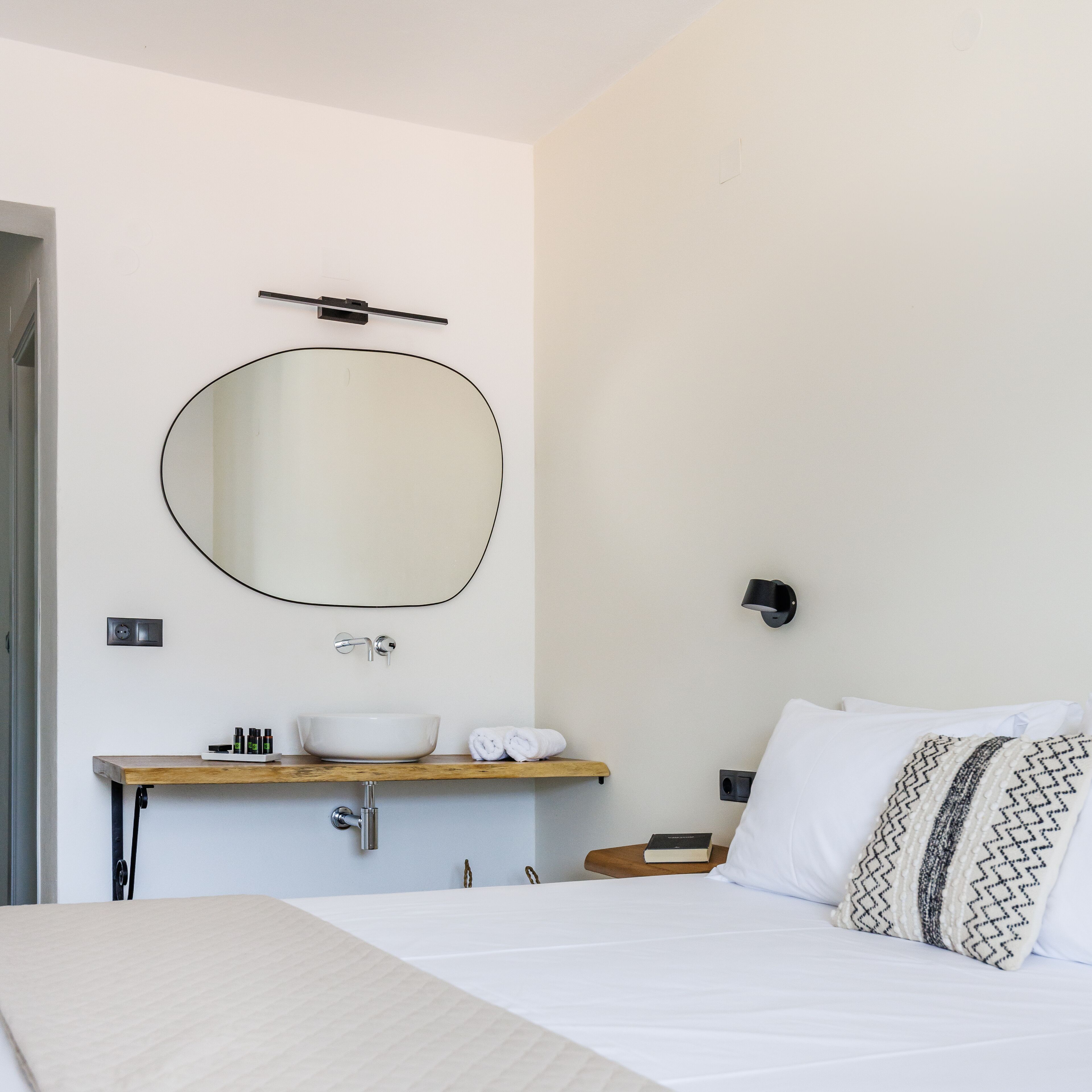 A hotel room with a bed, a bedside table with a book, a wall-mounted mirror, a sink, and a towel rack.