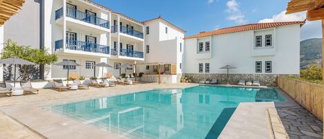 Una piscina con sillones de playa y sombrillas, rodeada de edificios blancos con balcones.