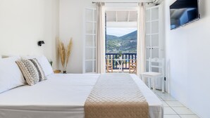 A bedroom with a bed, a TV, a balcony with a table and chair, and a view of a mountainous landscape.