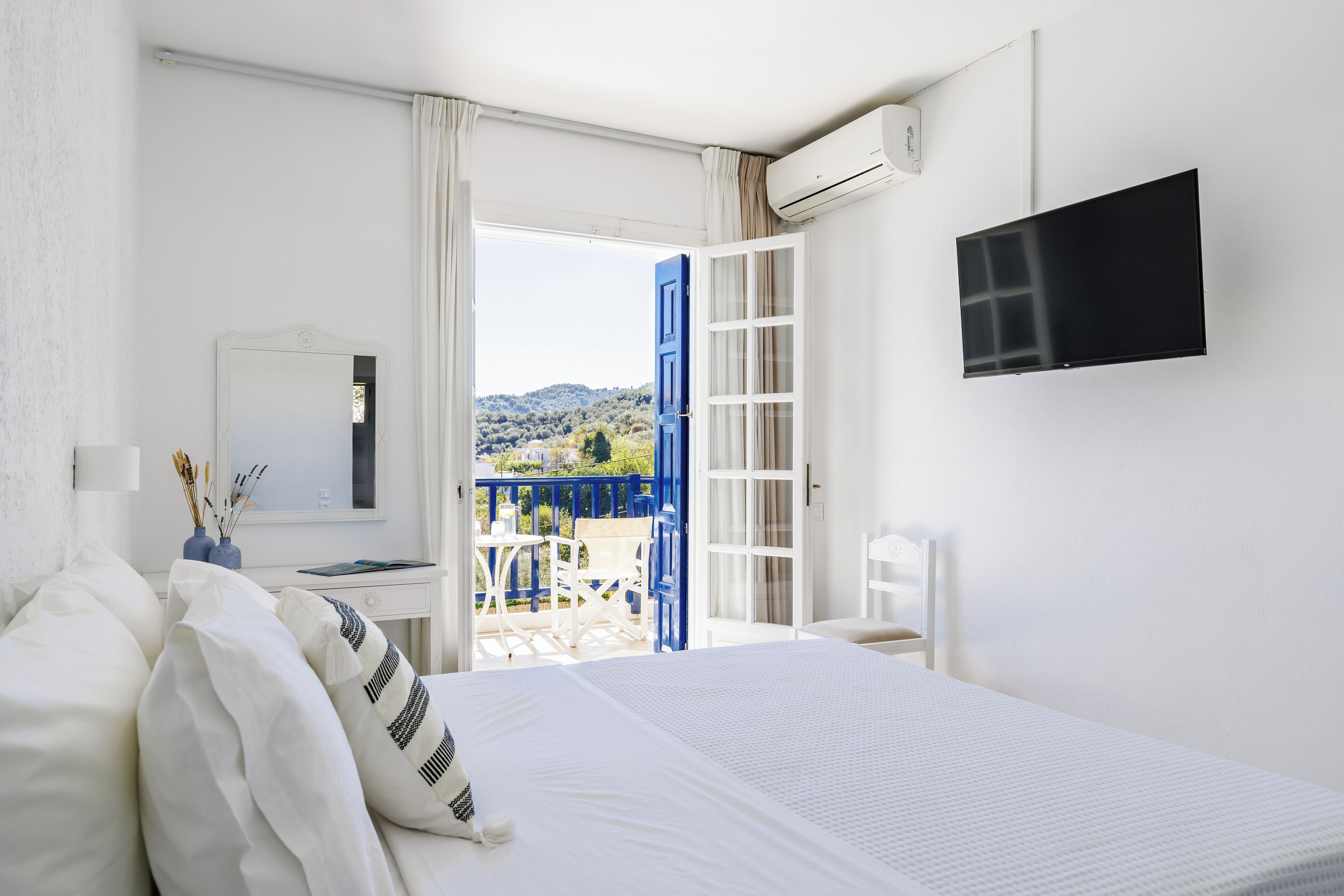 Habitación de hotel con cama, televisión, escritorio y vistas a un balcón con mobiliario exterior.