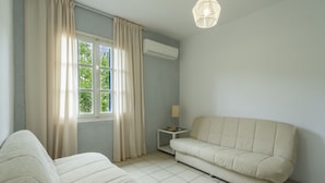 A room with two beds, a sofa, a small table with a lamp, and a window with curtains.
