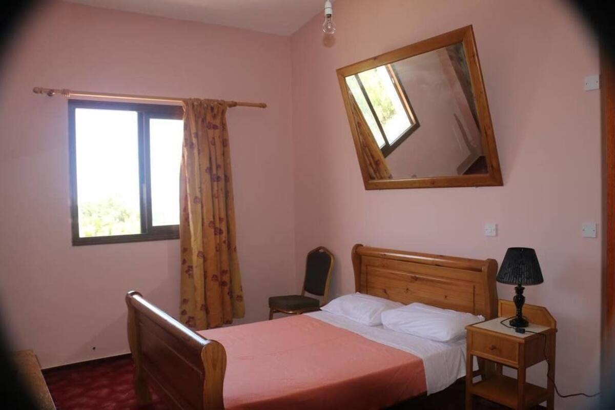 A bedroom with a bed, a nightstand, a chair, a window with curtains, and a mirror on the wall.