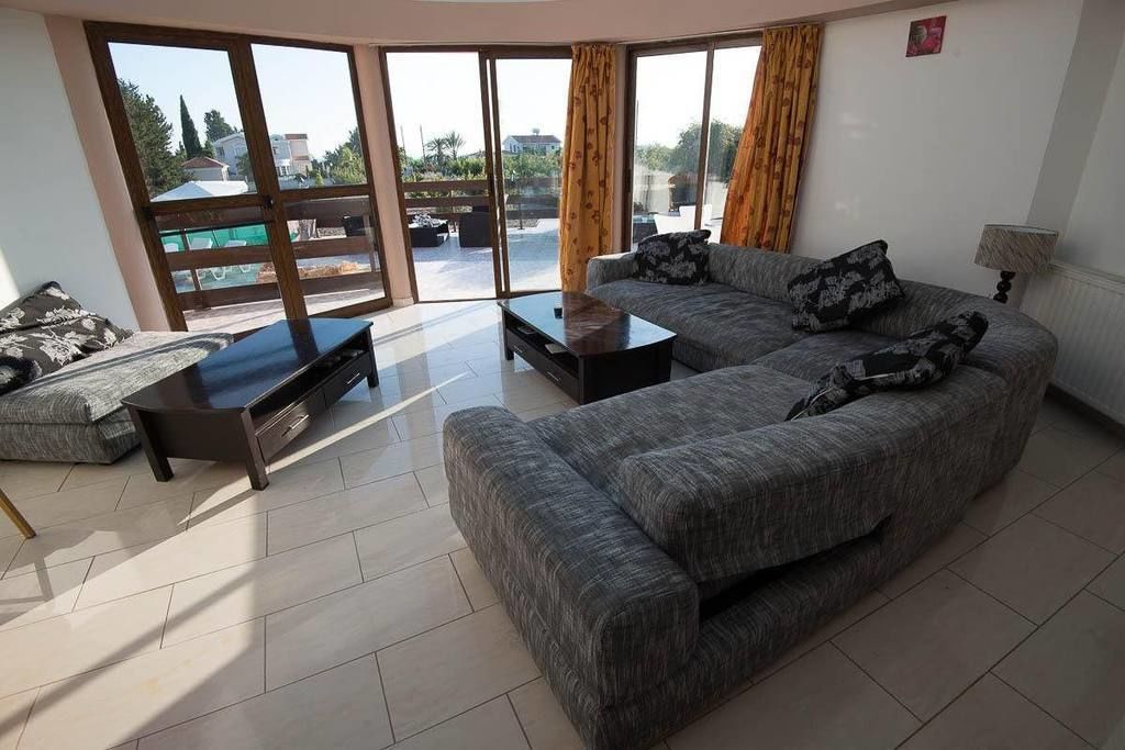 A spacious living room with a large sofa, a coffee table, and a TV. There are large windows with curtains, and a balcony is visible through the windows.