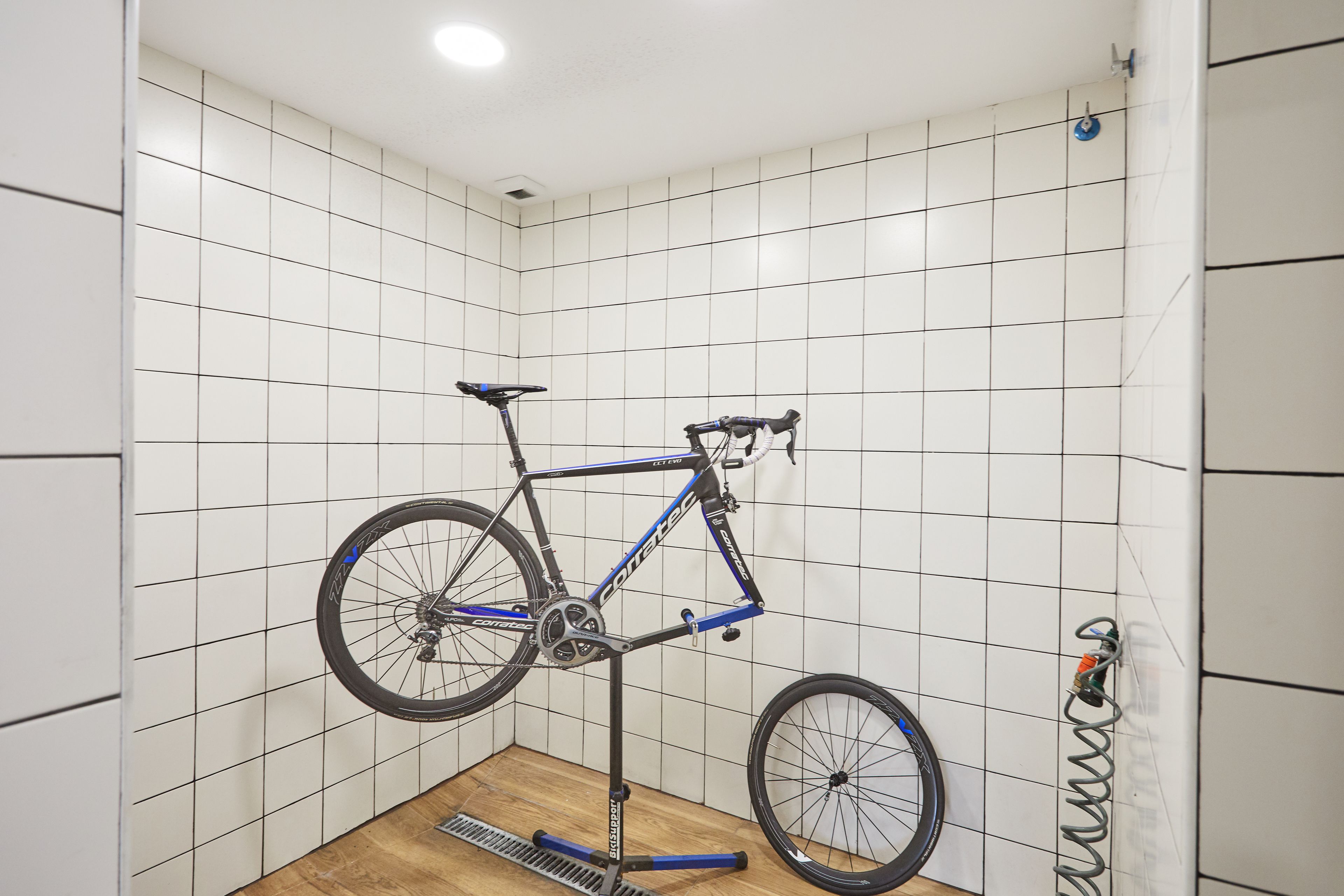 A bicycle mounted on a stand in a corner of a room with white tiled walls and a wooden floor.