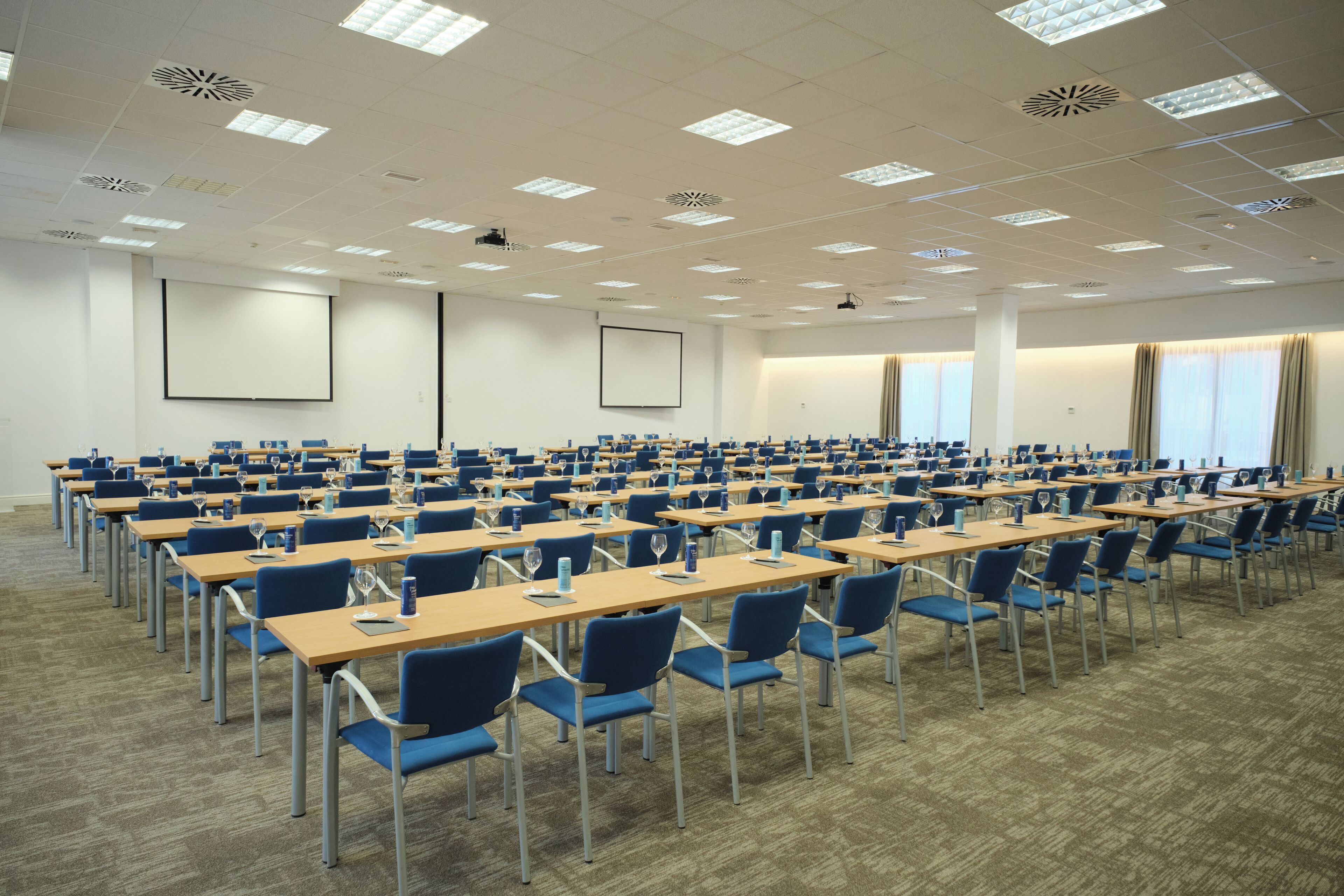 A conference room with rows of tables and chairs, large windows, and a projector screen.