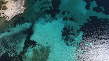 Luchtbeeld van een kustgebied met helder turquoise water, rotsformaties en schaarse begroeiing.